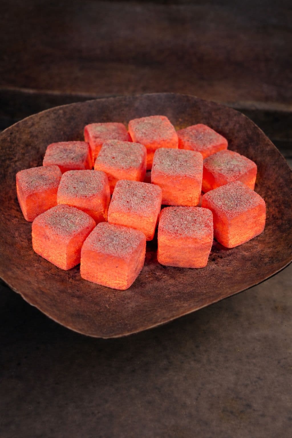 Glowing red-hot square charcoal cubes piled in a dark, rustic metal bowl.