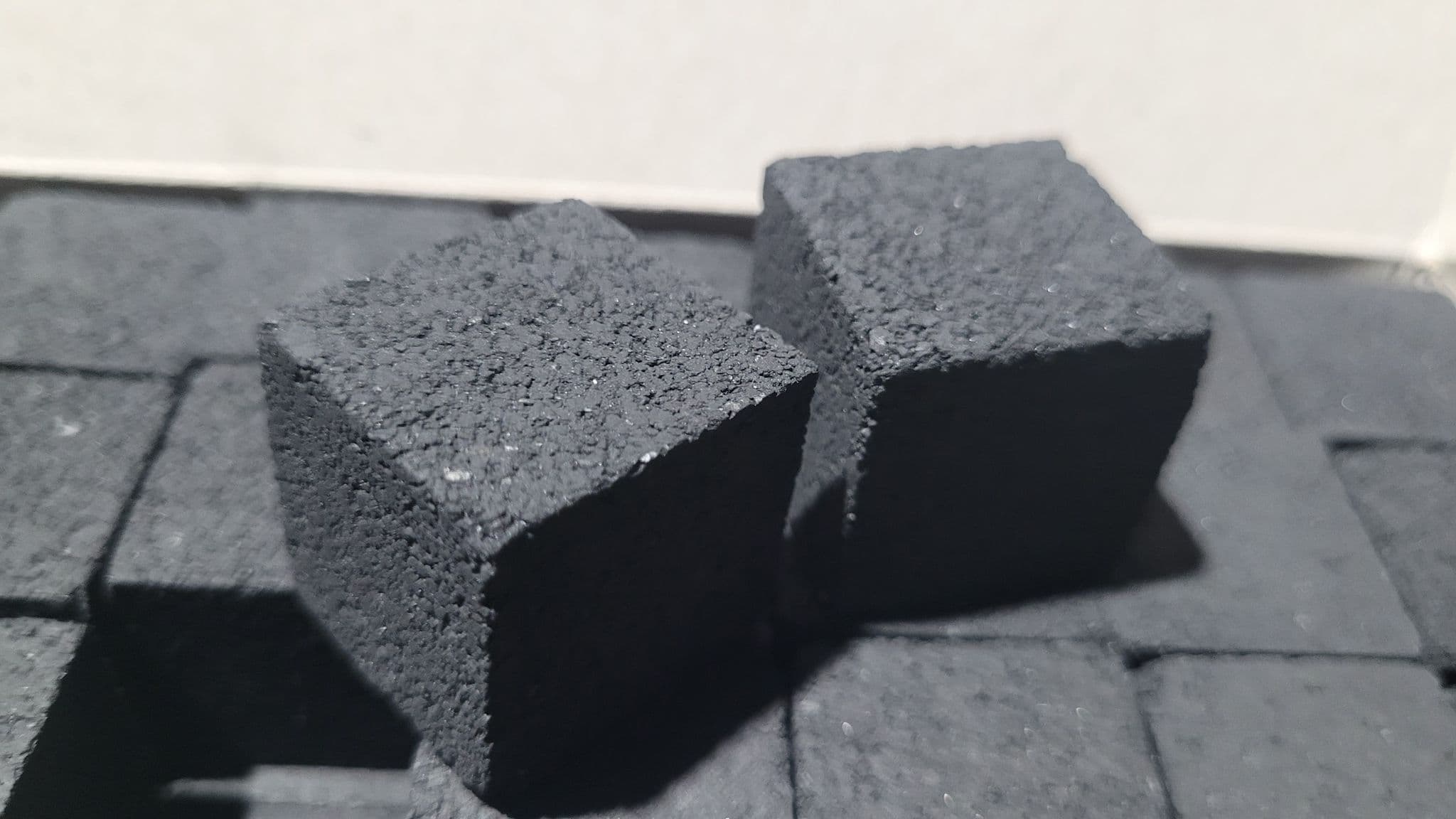 Two black, porous charcoal cubes with a rough texture resting on other charcoal pieces.