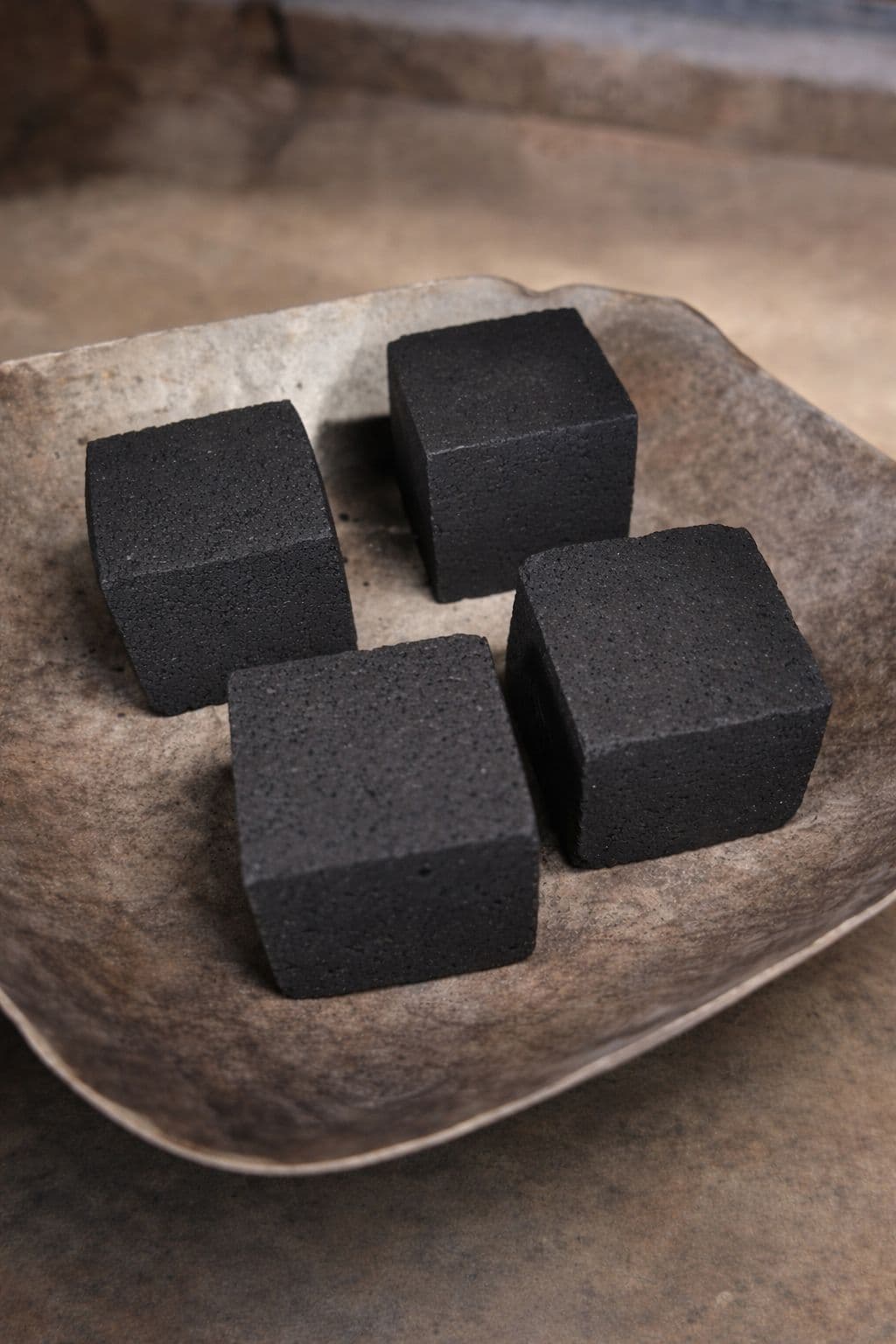Four black charcoal cubes arranged in a rustic, textured stone bowl on a neutral surface.